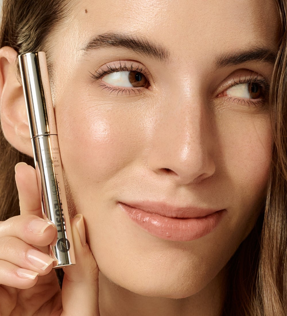 A woman holding a LashLush Serum tube beside her face.